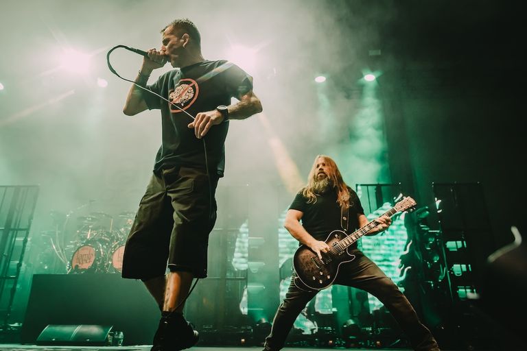 Lamb of God performing live with vocalist Randy Blythe singing into a microphone and guitarist Mark Morton playing on stage under green lights